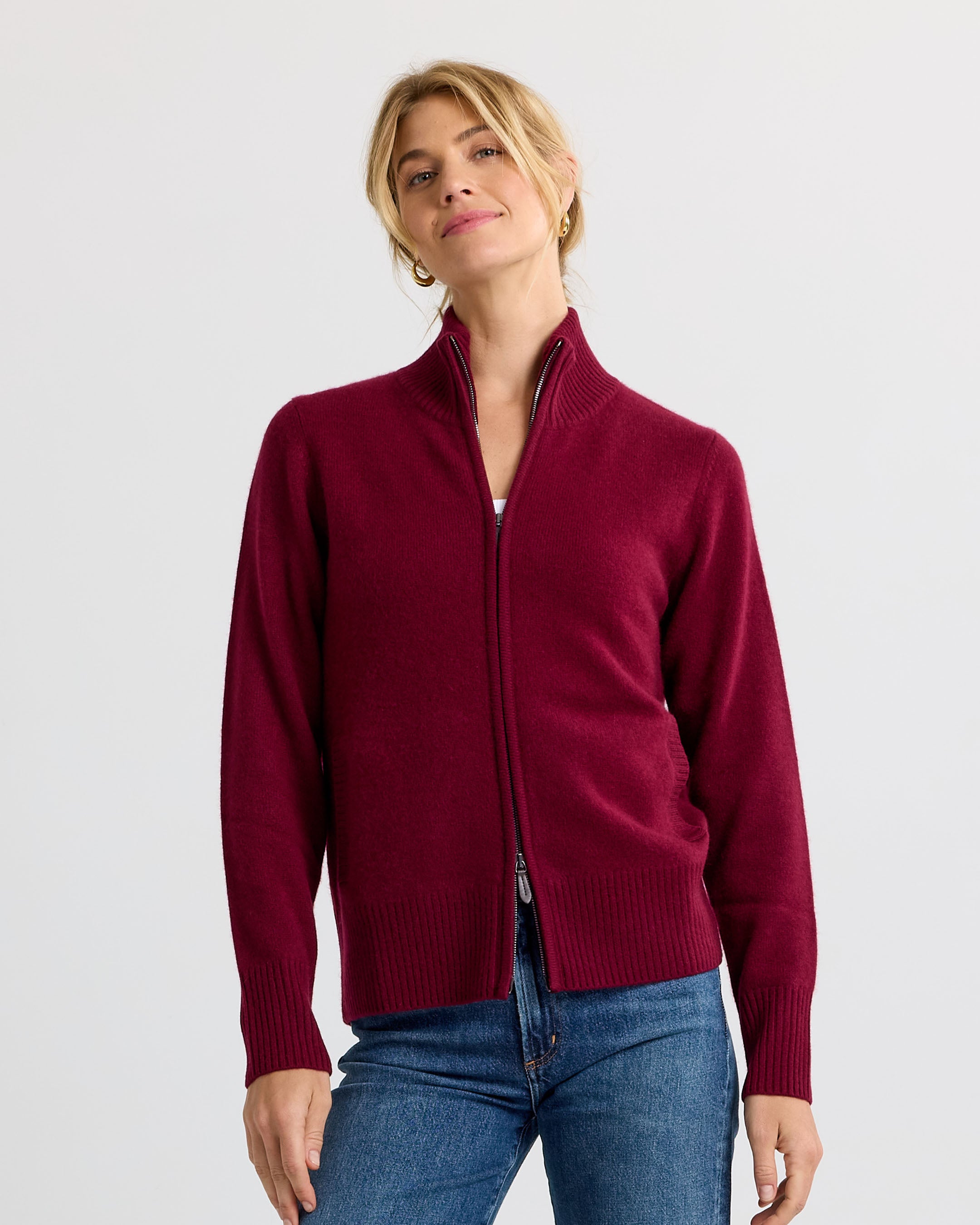 LIFESTYLE / EDITORIAL: Woman wearing a barolo-colored zip-up cashmere sweater and blue jeans against a plain background