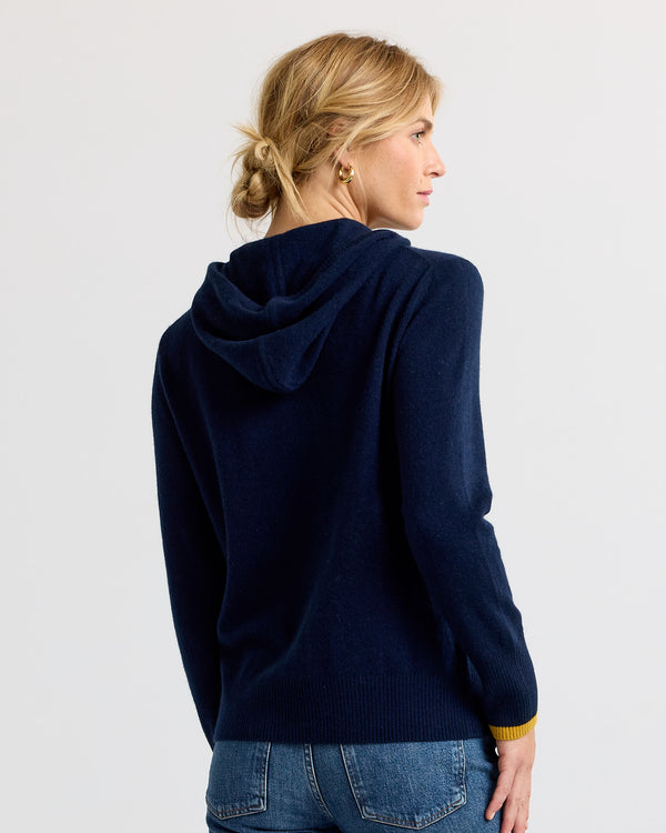 LIFESTYLE / EDITORIAL: Woman facing away wearing navy blue cashmere hoodie with mustard cuff detail and blue jeans