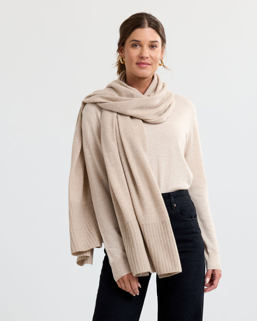 Three-quarter view image of a woman wearing beige cashmere sweater and scarf with black pants against white background