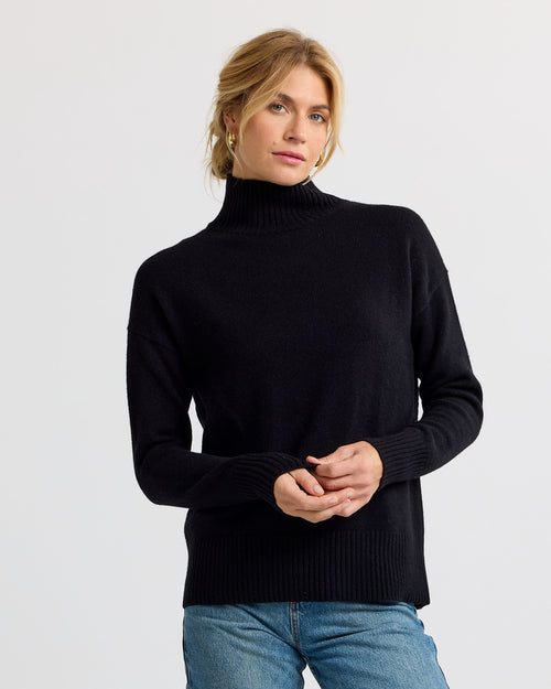 Black cashmere funnel neck sweater worn with blue jeans against a white background
