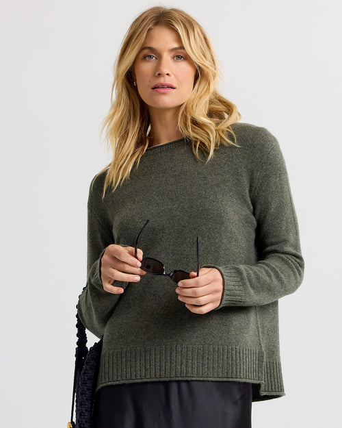 LIFESTYLE / EDITORIAL: Woman wearing dark olive cashmere sweater holding sunglasses against a plain background