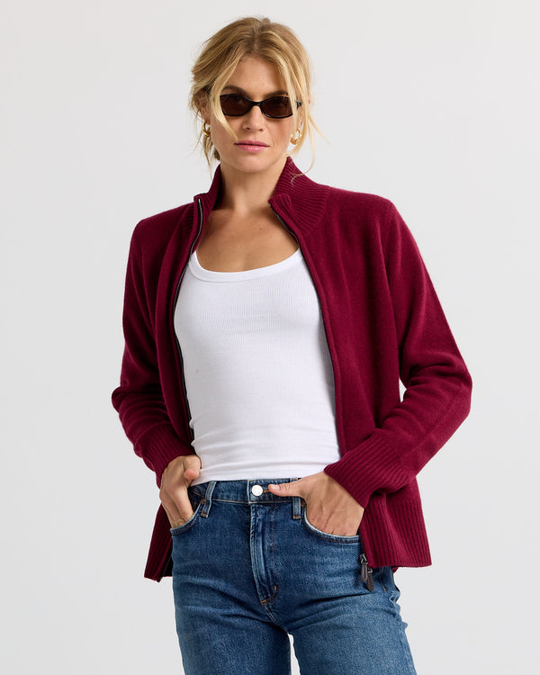 Woman wearing barolo-colored zip-up cashmere cardigan, white tank top, and blue jeans against white background