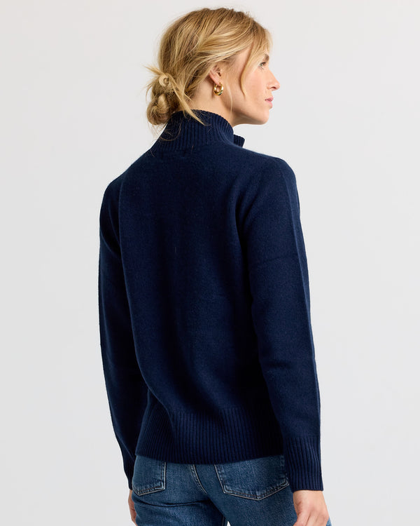 LIFESTYLE / EDITORIAL: Woman wearing navy cashmere turtleneck sweater, back view, hair in low bun, against plain background