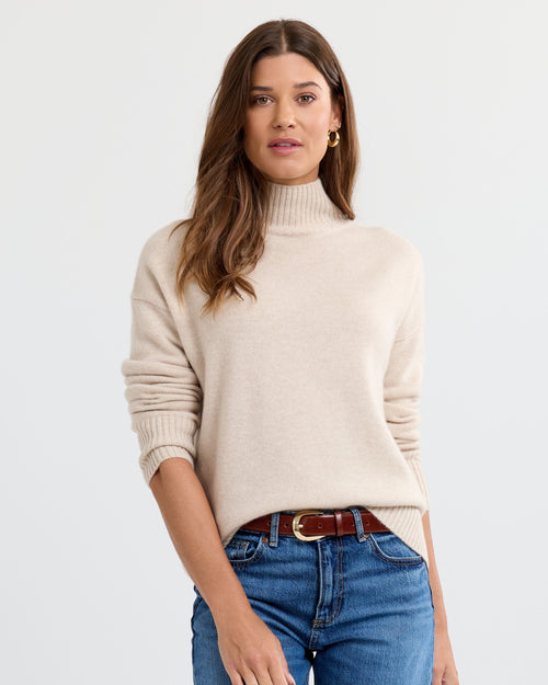 Oatmeal cashmere funnel neck sweater worn with blue jeans against a white background