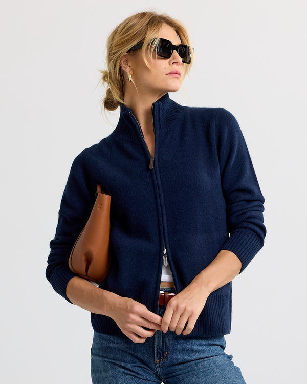 LIFESTYLE / EDITORIAL: Woman wearing navy zip-up cashmere sweater, sunglasses, holding brown leather bag, standing against