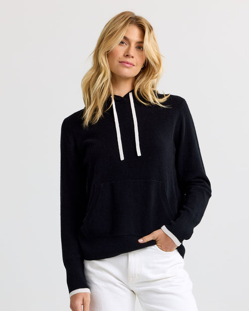 Front view image of a woman wearing a black cashmere hoodie with white drawstrings and white pants standing against a plain