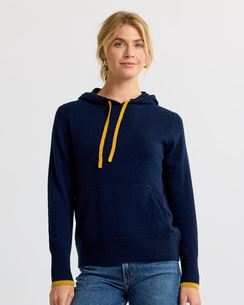 Woman wearing navy blue cashmere hoodie with yellow drawstrings and cuffs, paired with blue jeans, studio background