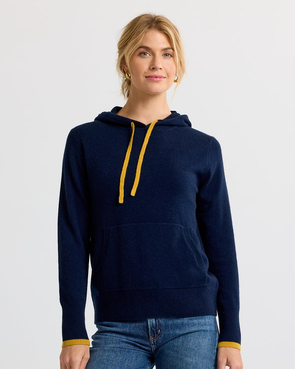 Woman wearing navy blue cashmere hoodie with yellow drawstrings and cuffs, paired with blue jeans, studio background