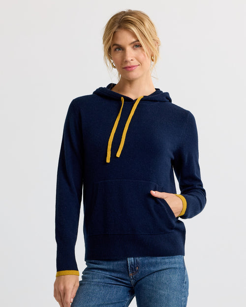 Woman wearing navy blue cashmere hoodie with yellow drawstrings and cuffs, paired with blue jeans against white background