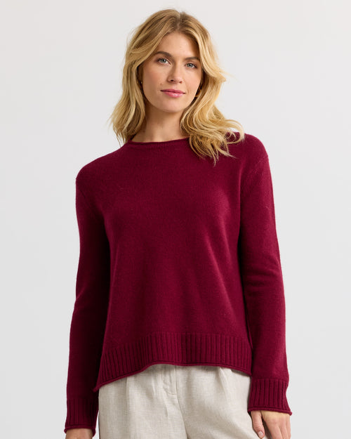 Woman wearing a barolo-colored cashmere sweater with ribbed cuffs and hem, paired with light beige pants against white