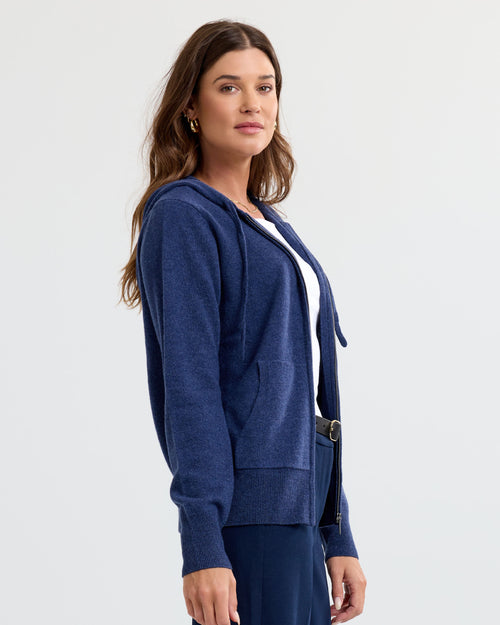 LIFESTYLE / EDITORIAL: Woman wearing indigo blue zip-up cashmere hoodie over white top, standing against plain background