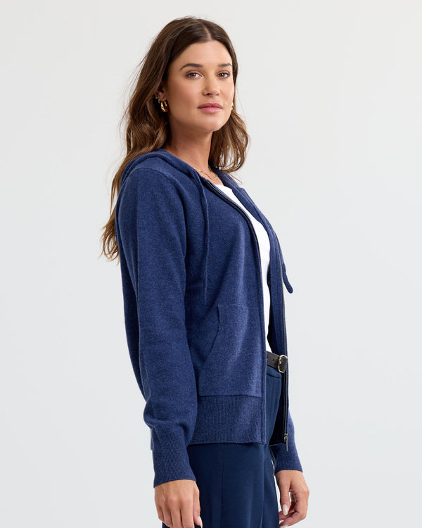 LIFESTYLE / EDITORIAL: Woman wearing indigo blue zip-up cashmere hoodie over white top, standing against plain background