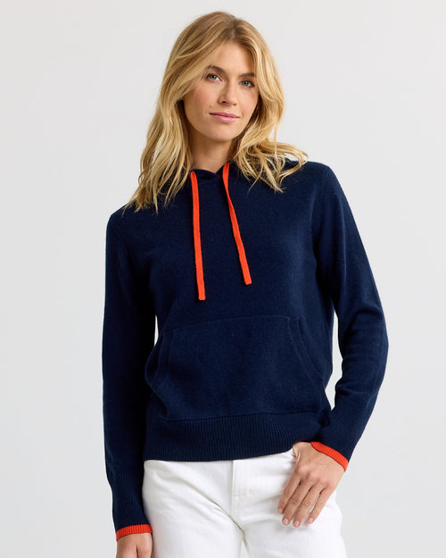 LIFESTYLE / EDITORIAL: Woman wearing navy cashmere hoodie with orange drawstrings and white pants against plain background