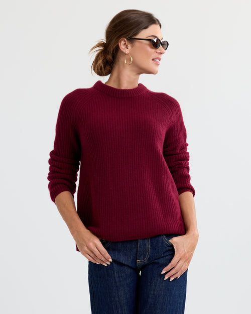 Woman wearing burgundy ribbed cashmere sweater and dark jeans, sunglasses, and gold hoop earrings against white background