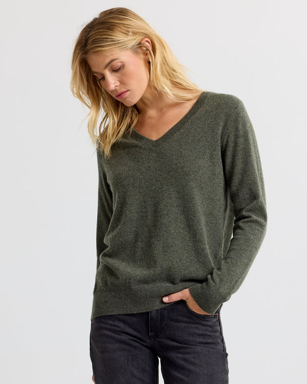 Woman wearing dark olive V-neck cashmere sweater and black jeans, standing against plain light background