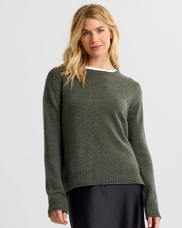 Woman wearing dark olive cashmere sweater with ribbed cuffs and hem, standing against a plain light background