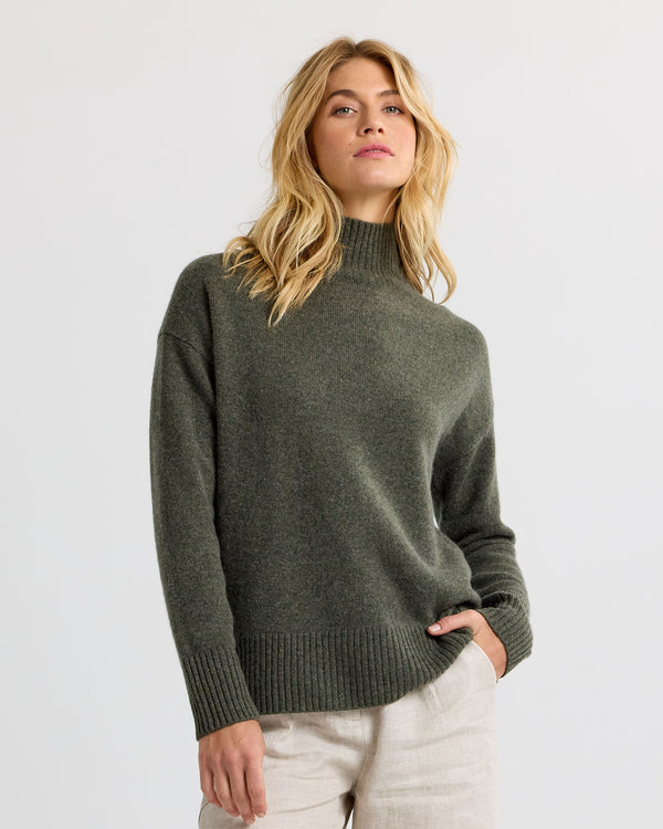 Dark olive cashmere funnel neck sweater with light trousers against a plain background