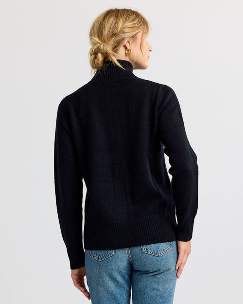 Woman wearing black turtleneck cashmere sweater and blue jeans, viewed from the back against a plain background