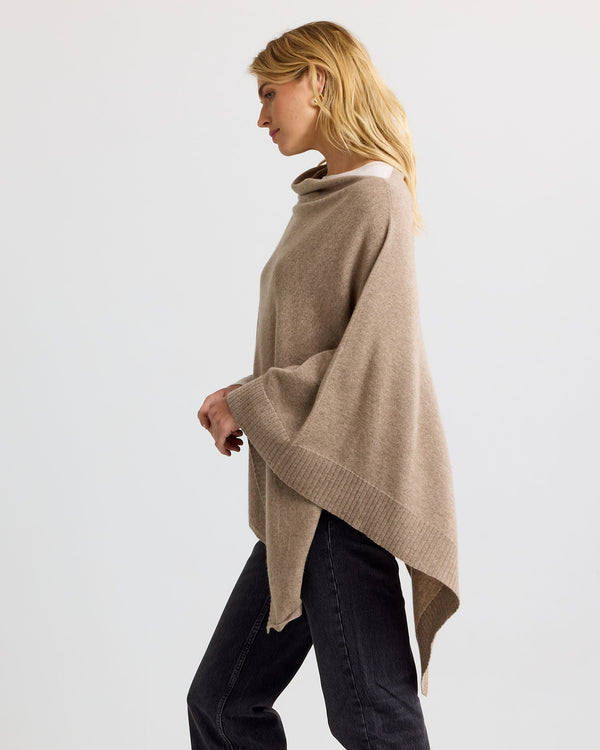 LIFESTYLE / EDITORIAL: Woman in beige cashmere poncho and black jeans, side profile against plain background