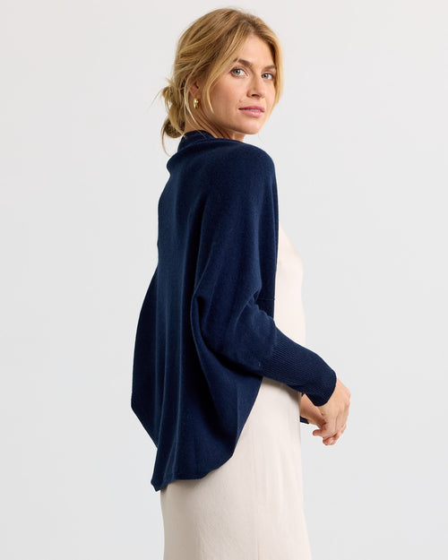 LIFESTYLE / EDITORIAL: Woman wearing navy cashmere poncho over cream dress, looking over shoulder against white background