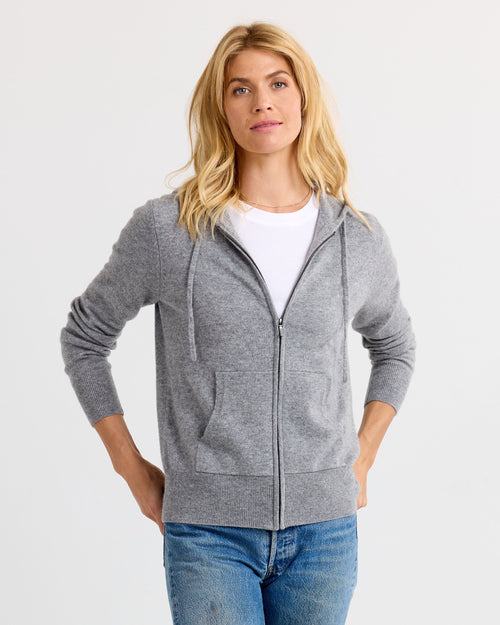Woman wearing grey zip-up cashmere hoodie over white tee and blue jeans, standing against white background