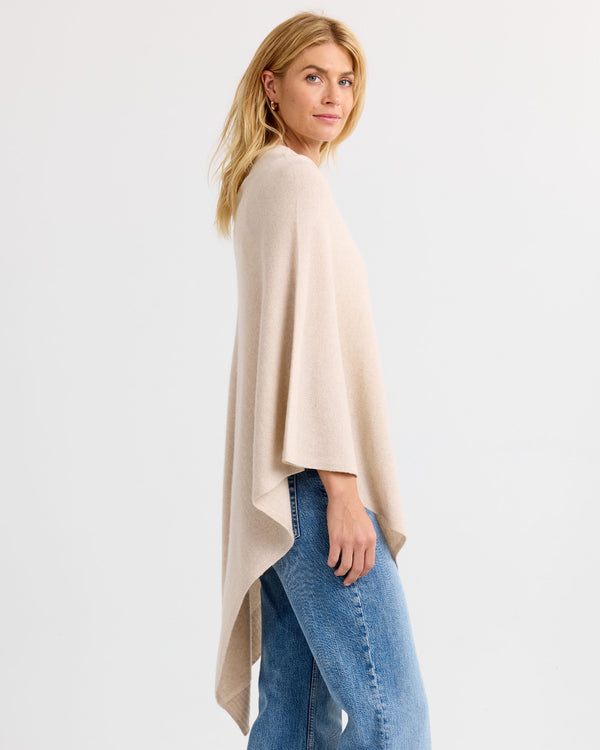 Woman wearing oatmeal-colored cashmere poncho with blue jeans, standing sideways against a white background