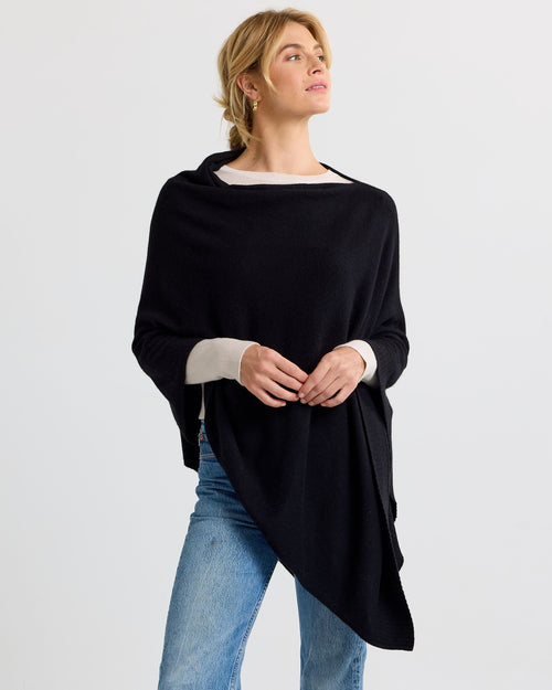 LIFESTYLE / EDITORIAL: Woman wearing black cashmere poncho over light sweater and jeans, standing against plain background