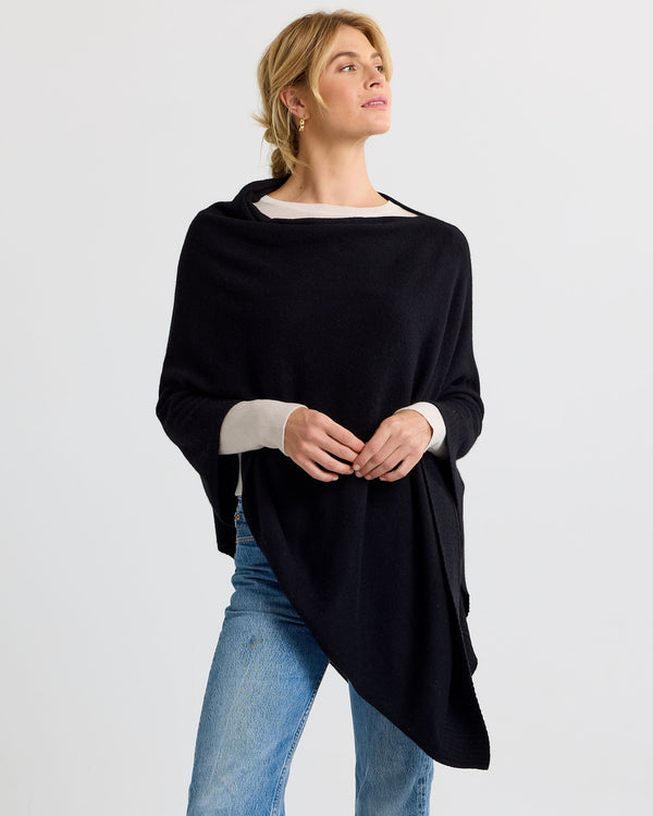 LIFESTYLE / EDITORIAL: Woman wearing black cashmere poncho over light sweater and jeans, standing against plain background