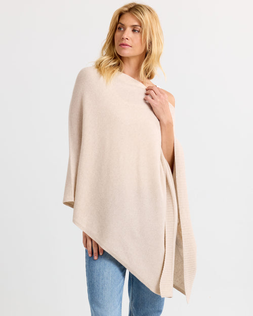 Three-quarter view image of a woman wearing an oatmeal cashmere poncho with blue jeans against a white background