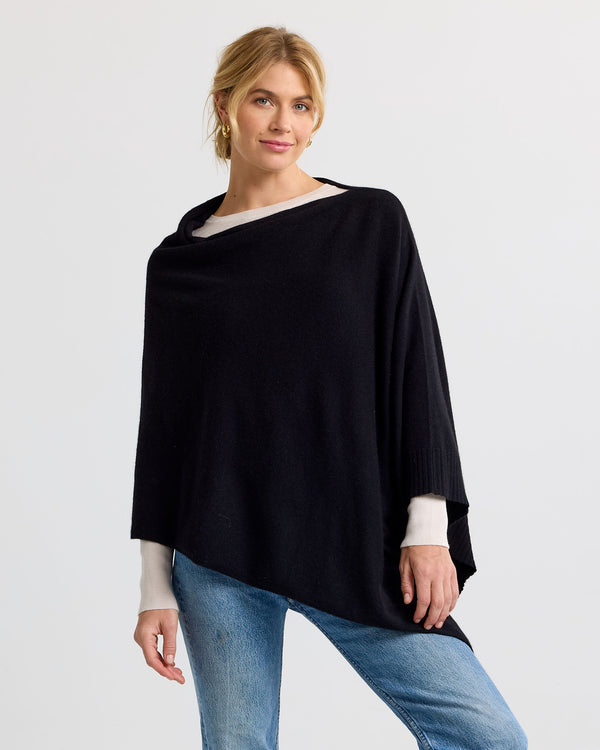 Three-quarter view image of a woman wearing a black asymmetrical cashmere poncho over white sleeves and blue jeans
