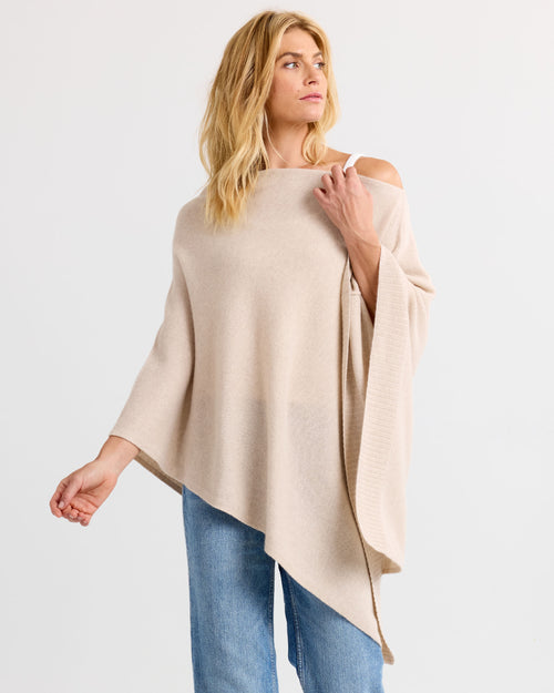 Three-quarter view image of a woman wearing an oatmeal cashmere poncho with blue jeans against a white background