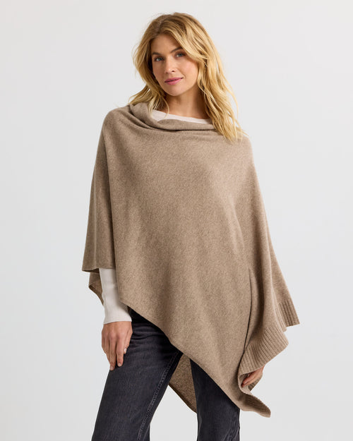 LIFESTYLE / EDITORIAL: Woman wearing a driftwood-colored cashmere poncho over white top and dark jeans against white