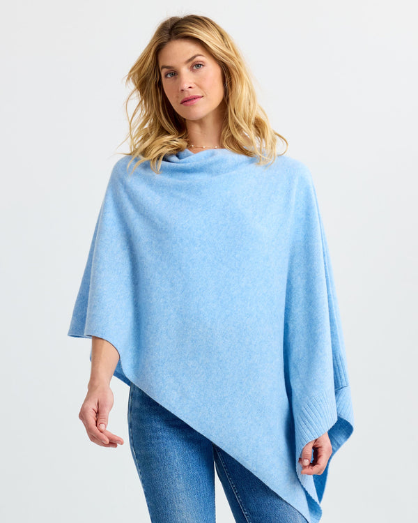 LIFESTYLE / EDITORIAL: Woman wearing light blue asymmetrical cashmere poncho and jeans against plain background