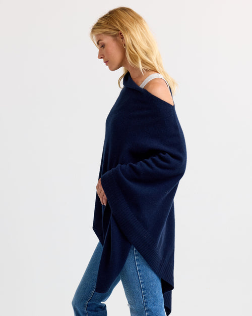 LIFESTYLE / EDITORIAL: Woman wearing navy cashmere poncho over white top and jeans, side profile against plain background