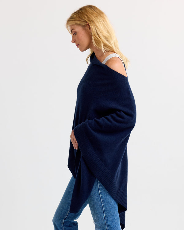LIFESTYLE / EDITORIAL: Woman wearing navy cashmere poncho over white top and jeans, side profile against plain background