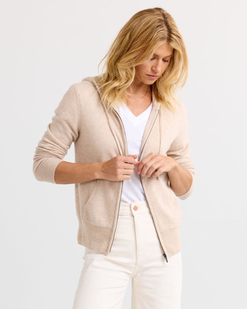 Woman wearing oatmeal zip-up cashmere hoodie over white V-neck shirt and white pants against plain background