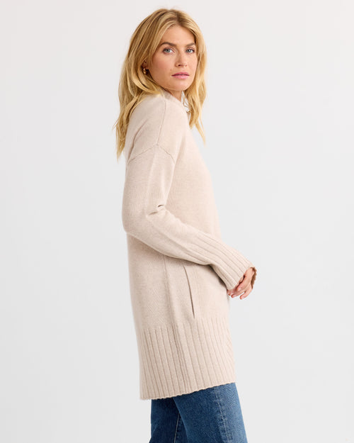 LIFESTYLE / EDITORIAL: Woman wearing long oatmeal cashmere sweater and blue jeans, standing sideways against white background