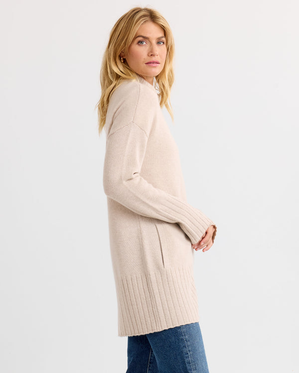 LIFESTYLE / EDITORIAL: Woman wearing long oatmeal cashmere sweater and blue jeans, standing sideways against white background