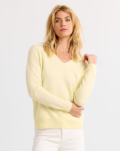 Three-quarter image of a woman wearing a light yellow V-neck cashmere sweater and white pants with hand raised
