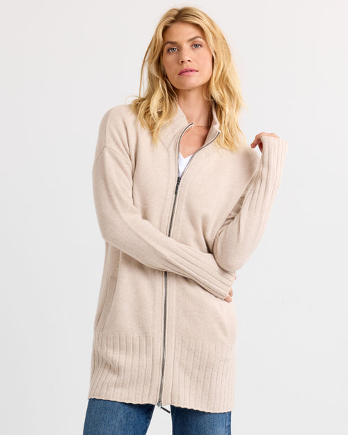 LIFESTYLE / EDITORIAL: Woman wearing long oatmeal zip-up cashmere sweater with ribbed cuffs and hem, standing against white