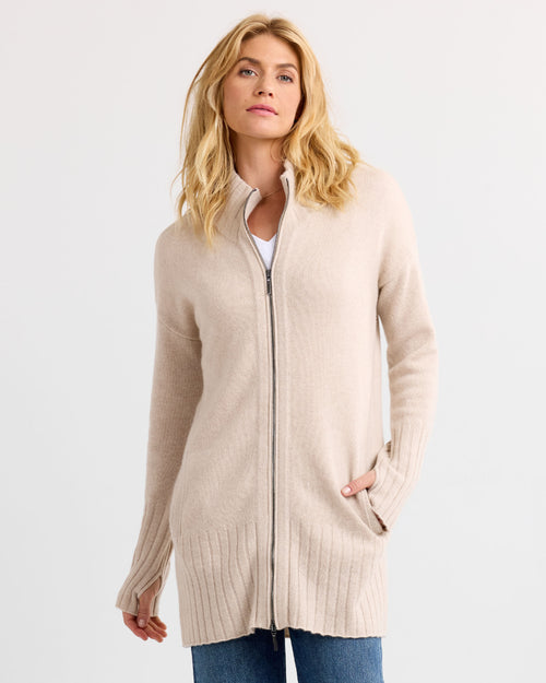 LIFESTYLE / EDITORIAL: Woman wearing long oatmeal zip-up cashmere sweater with thumb holes, standing against white background