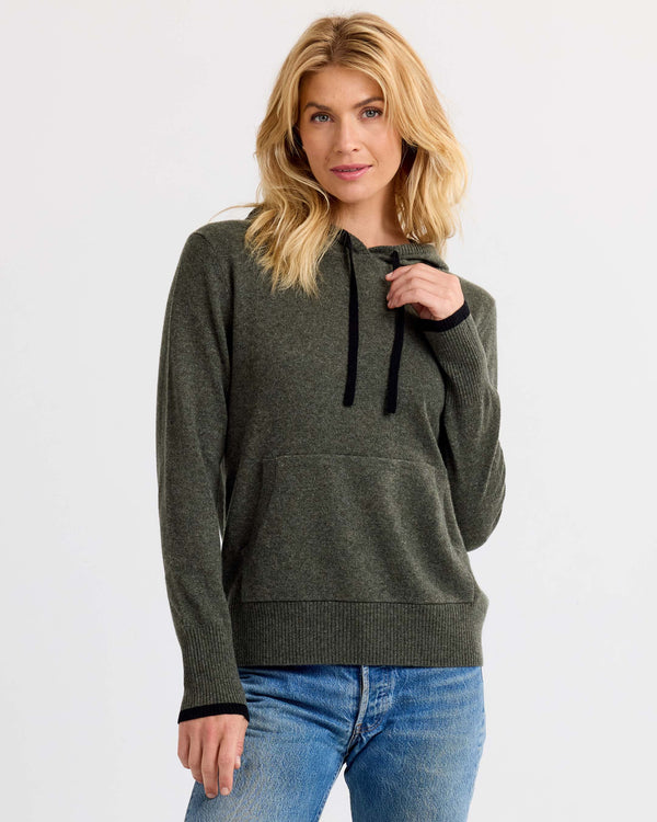 Woman wearing dark olive and black cashmere hoodie with front pocket and blue jeans against white background