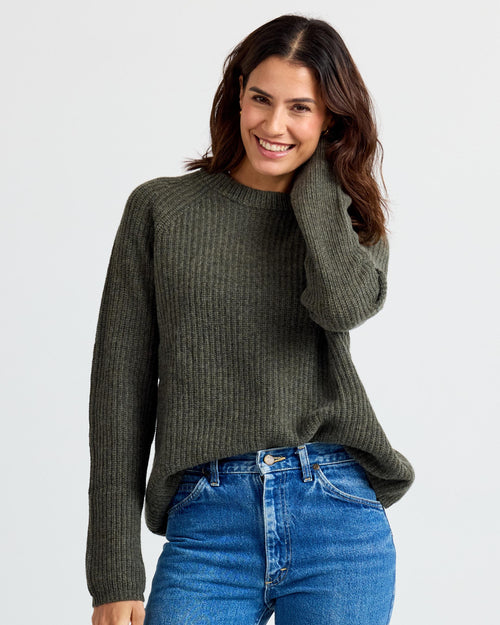 LIFESTYLE / EDITORIAL: Smiling woman wearing dark olive ribbed cashmere sweater and blue jeans against white background