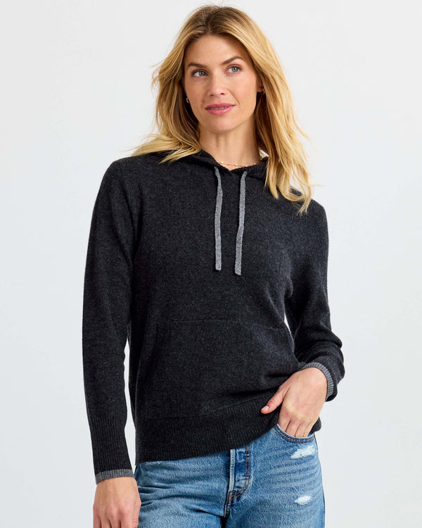 Woman wearing charcoal gray cashmere hoodie sweater with light gray drawstrings and blue jeans against white background