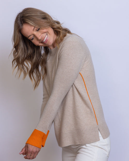 LIFESTYLE / EDITORIAL: Woman smiling, wearing beige cashmere sweater with orange cuffs and white pants against gray
