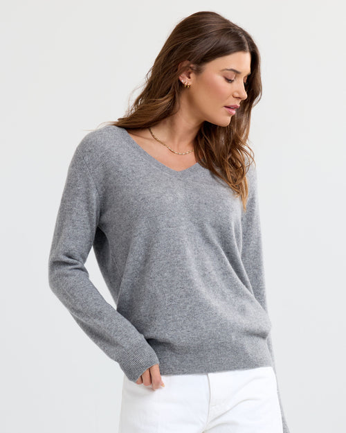 LIFESTYLE / EDITORIAL: Woman wearing grey V-neck cashmere sweater and white pants, posing against a plain background