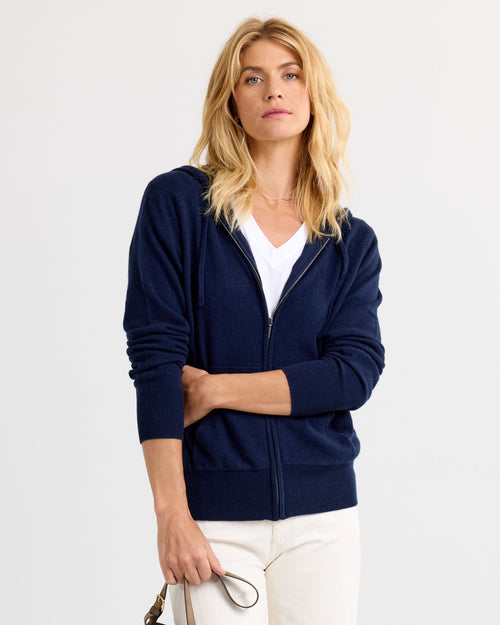 LIFESTYLE / EDITORIAL: Woman wearing navy zip-up cashmere hoodie over white shirt, standing against plain background