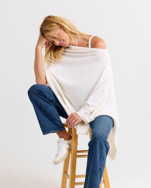 LIFESTYLE / EDITORIAL: Woman sitting on wooden stool wearing ivory off-shoulder cashmere sweater and blue jeans