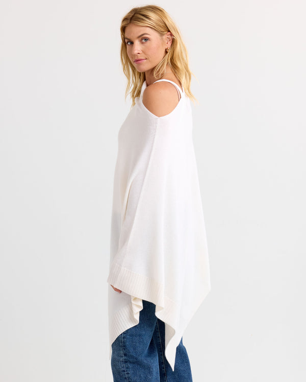 LIFESTYLE / EDITORIAL: Woman wearing ivory cold-shoulder cashmere poncho with jeans, standing sideways against white