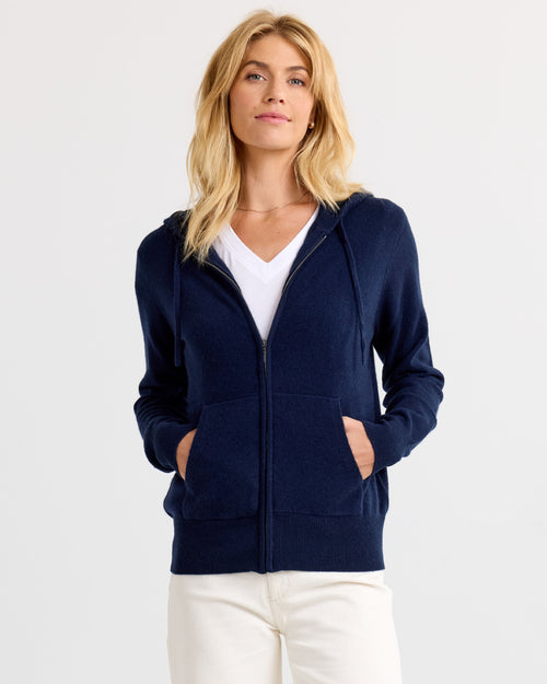 LIFESTYLE / EDITORIAL: Woman wearing navy zip-up cashmere hoodie with white shirt and cream pants against white background
